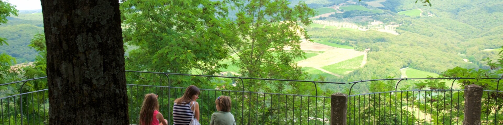 Três pessoas sentadas num banco, com vista para um vale com cerca e árvores.
