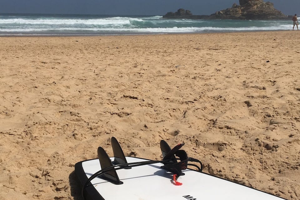 Una tabla de surf blanca con aletas y una correa roja sobre una playa arenosa, con olas al fondo y una formación rocosa.