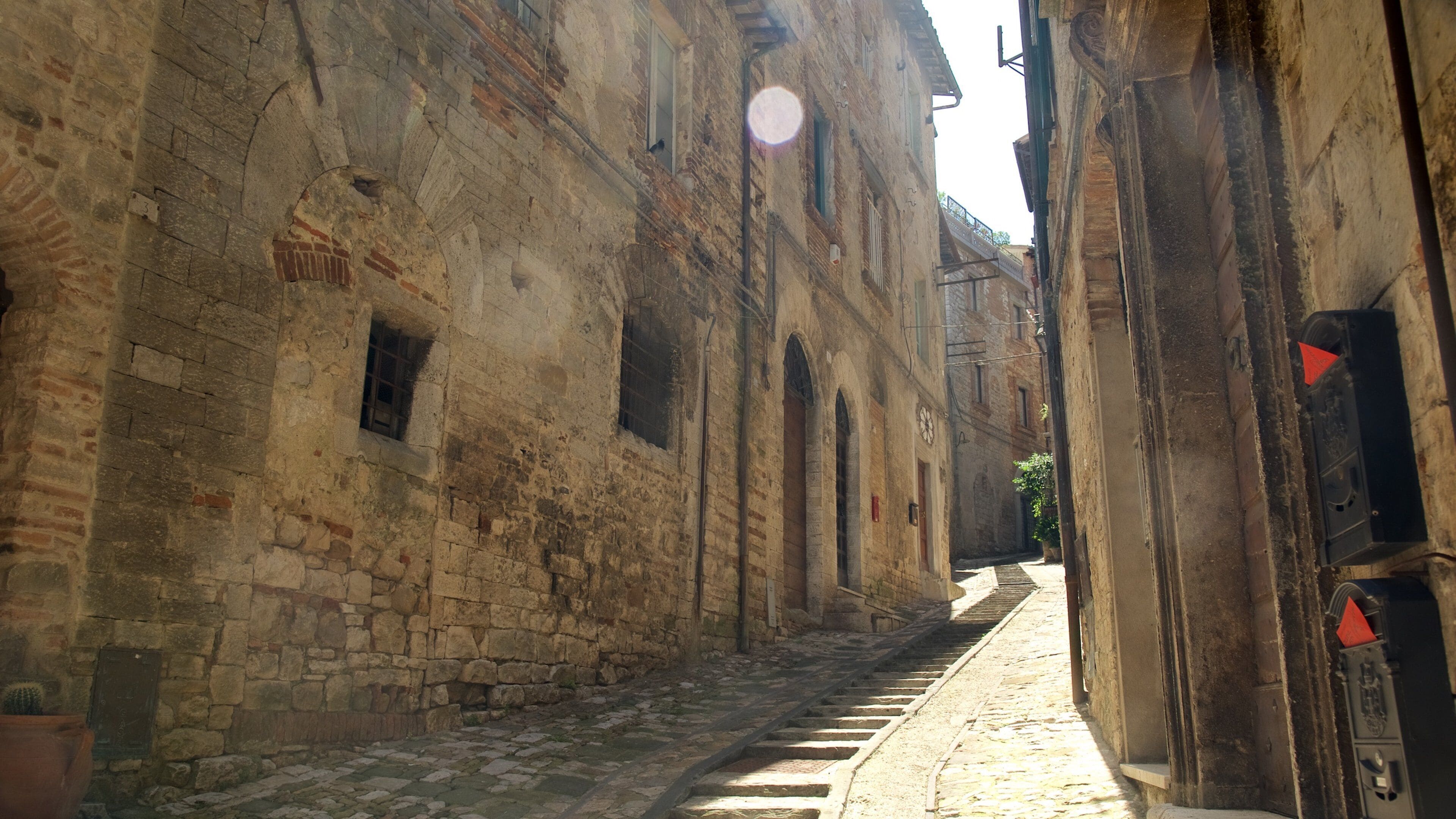 Uma rua de paralelepípedos estreita, ladeada por edifícios antigos de pedra com janelas em arco e um relógio.