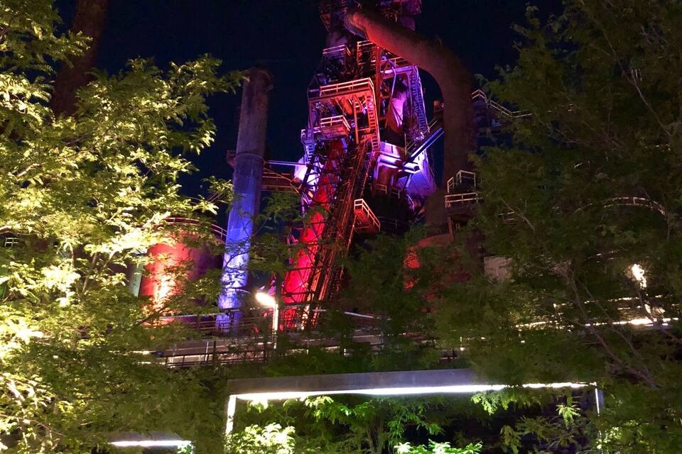 A nighttime view of a large industrial structure with scaffolding, surrounded by trees and illuminated by artificial lights.
