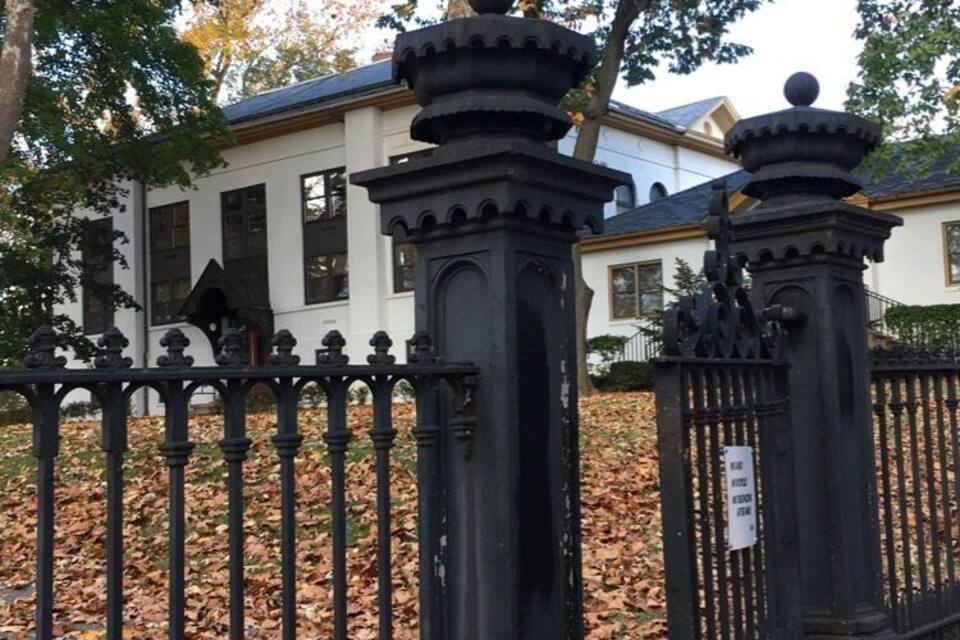 A black wrought iron fence with ornate pillars, a white building with black shutters, and autumn leaves on the ground.