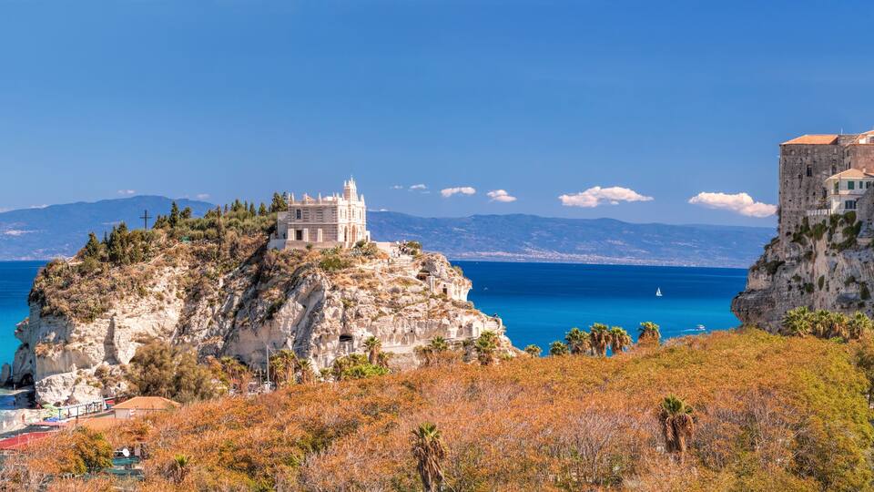 A coastal town with historic buildings, a prominent church on a cliff, and a clear blue sea.