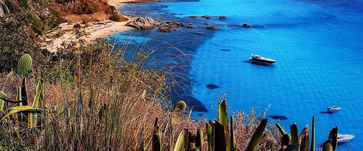 A coastal landscape with a clear blue sea, a sandy beach, rocky outcrops, and a mountainous backdrop.
