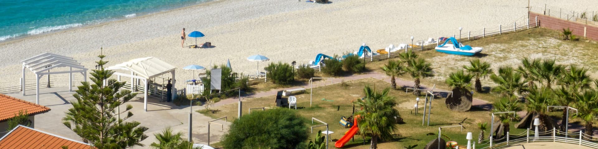 Aerial view of a wide sandy beach meeting a bright turquoise sea. Scattered colorful umbrellas and small white structures dot the shoreline. It is Zambrone beach, near Tropea, in Calabria, Italy.