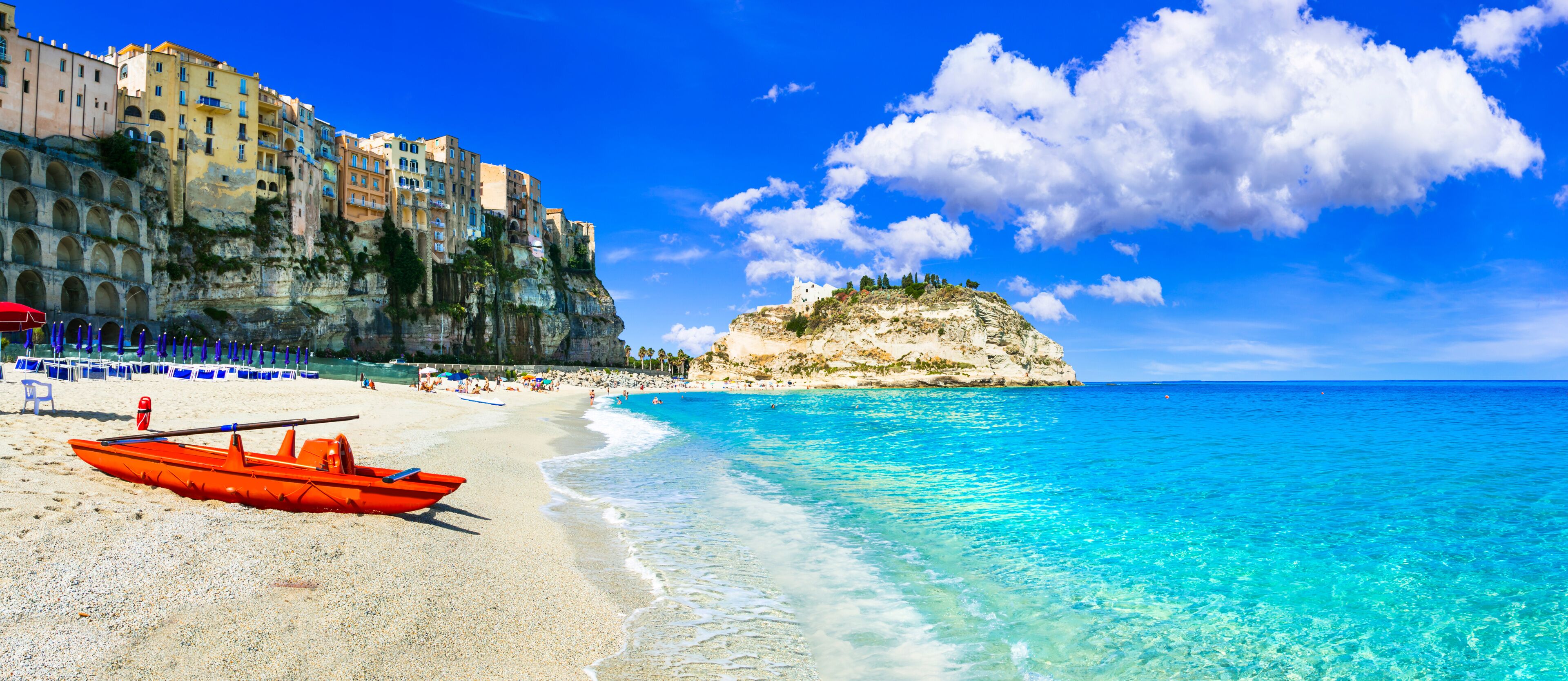 Ein Strand mit klarem türkisblauem Wasser, einem Felsen mit Gebäuden und einem roten Boot im Sand.