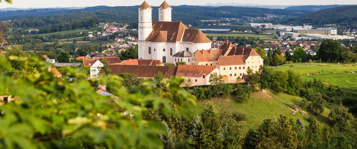 A historic white church with two towers, surrounded by lush greenery and a town in the background.