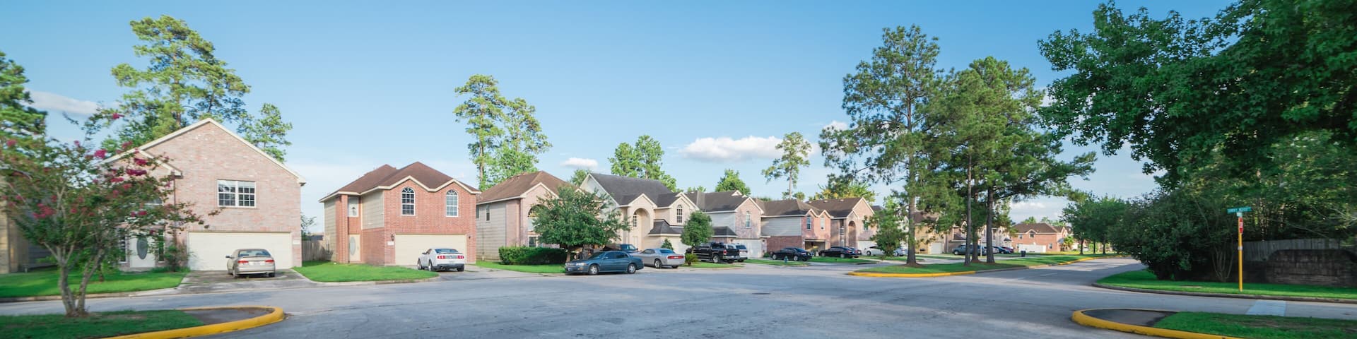 Suburban residential area, row of modern townhomes in Humble, Texas, US. Red brick houses surrounded with tall pine trees, cloud blue sky. Panorama view street intersection and multi-story townhouses.