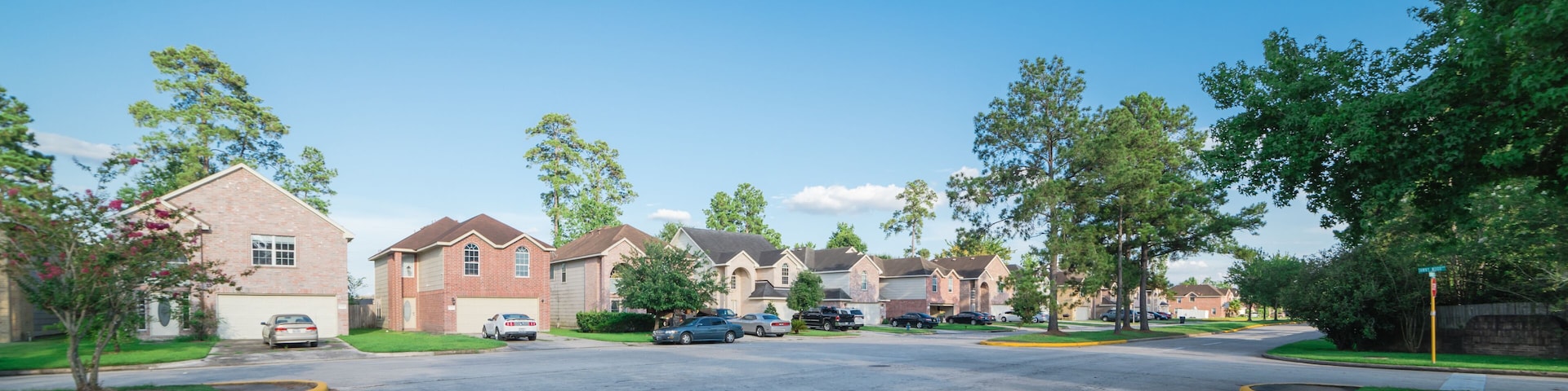 Suburban residential area, row of modern townhomes in Humble, Texas, US. Red brick houses surrounded with tall pine trees, cloud blue sky. Panorama view street intersection and multi-story townhouses.