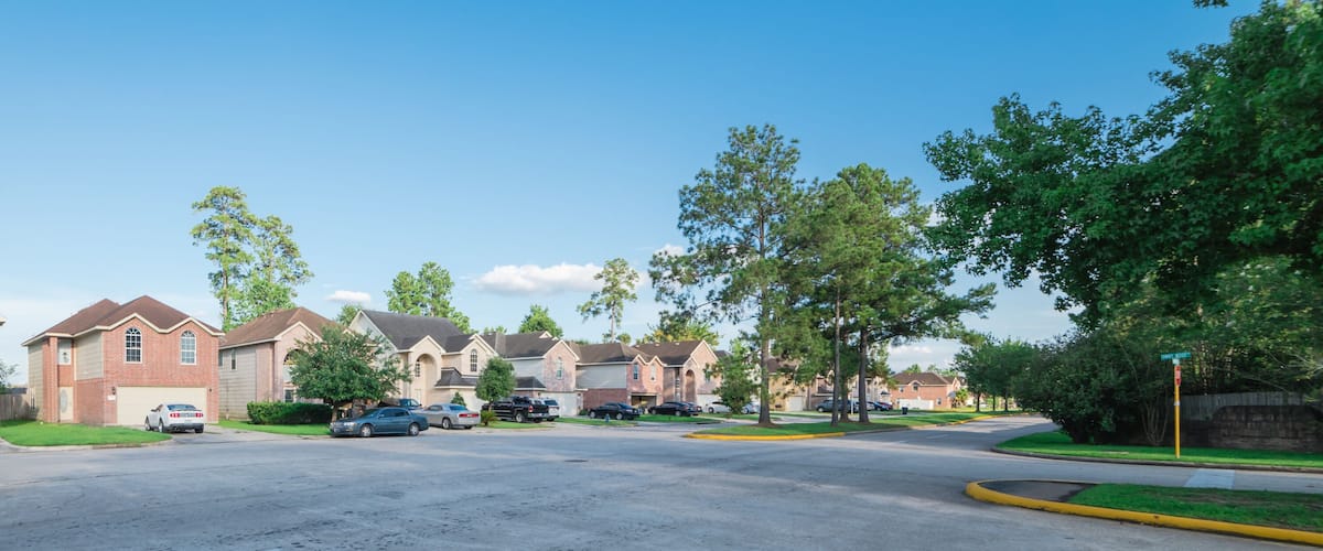 A suburban street lined with brick houses, parked cars, and trees.