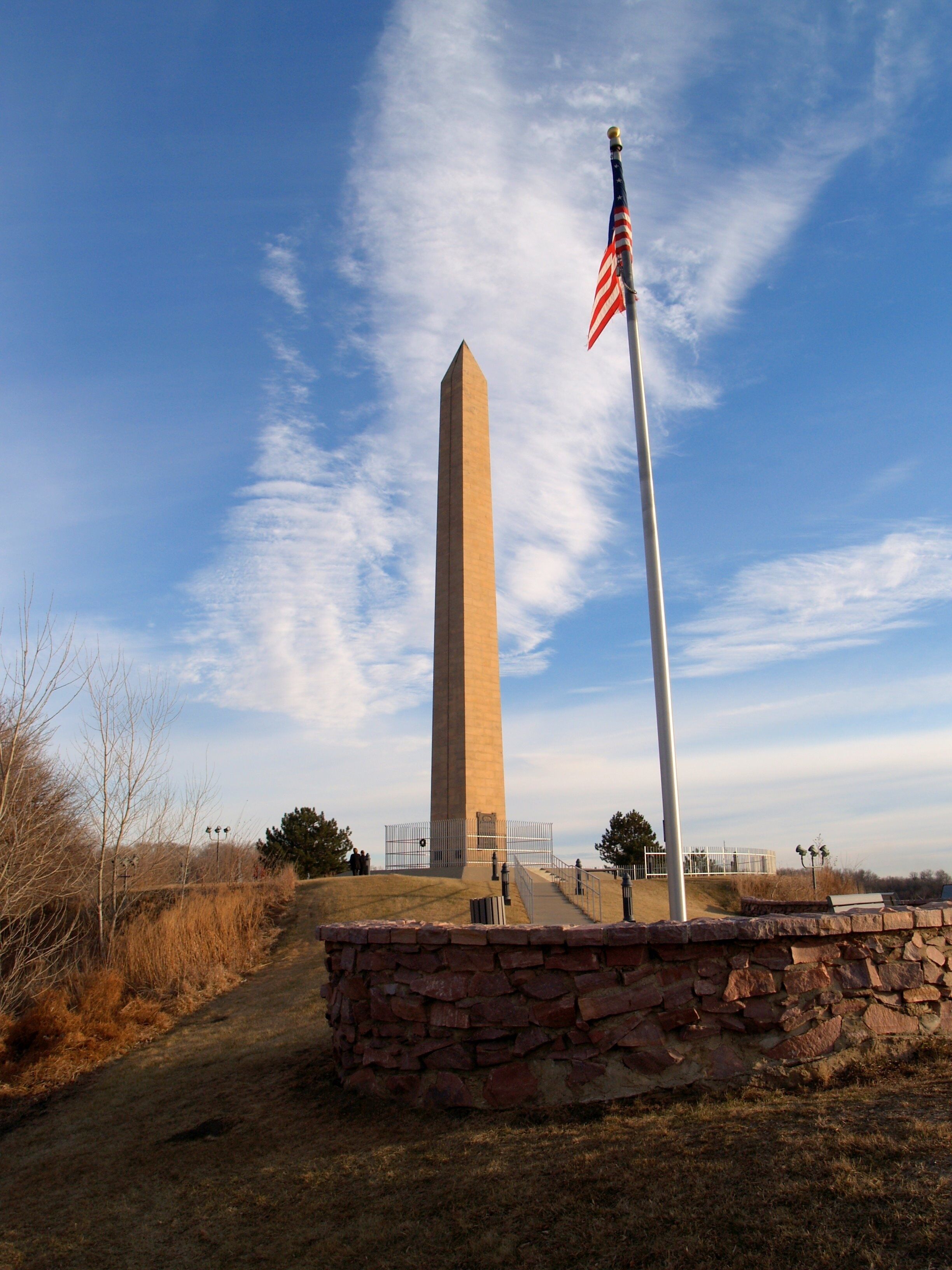 A tall obelisk with an American flag, surrounded by a stone wall and a clear sky.