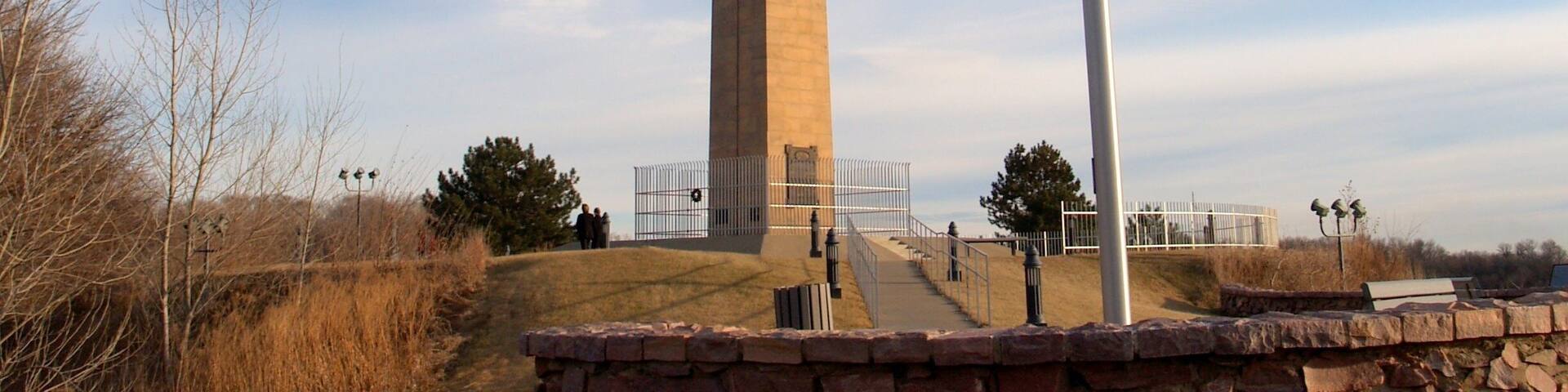 A tall obelisk with an American flag, surrounded by a stone wall and a clear sky.