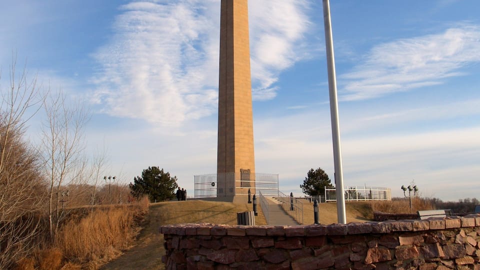 A tall obelisk with an American flag, surrounded by a stone wall and a clear sky.