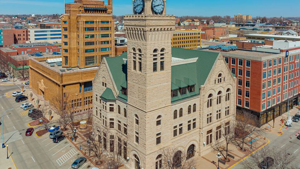 A historic building with a clock tower, surrounded by modern architecture and a parking lot.