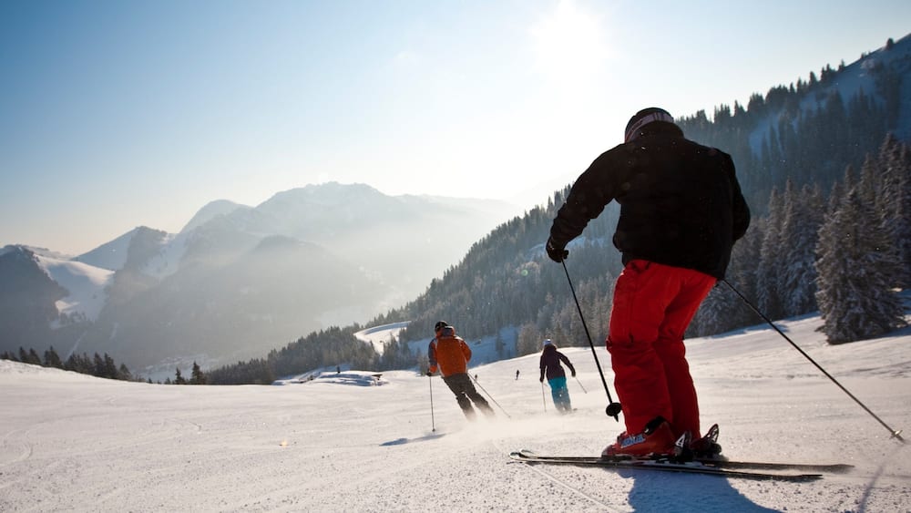 Skiers on a snowy mountain slope with a clear sky and forested hills in the background.