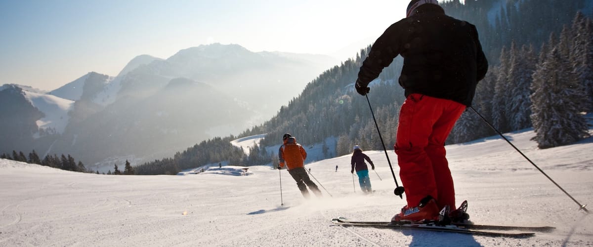 Skiers on a snowy mountain slope with a clear sky and forested hills in the background.