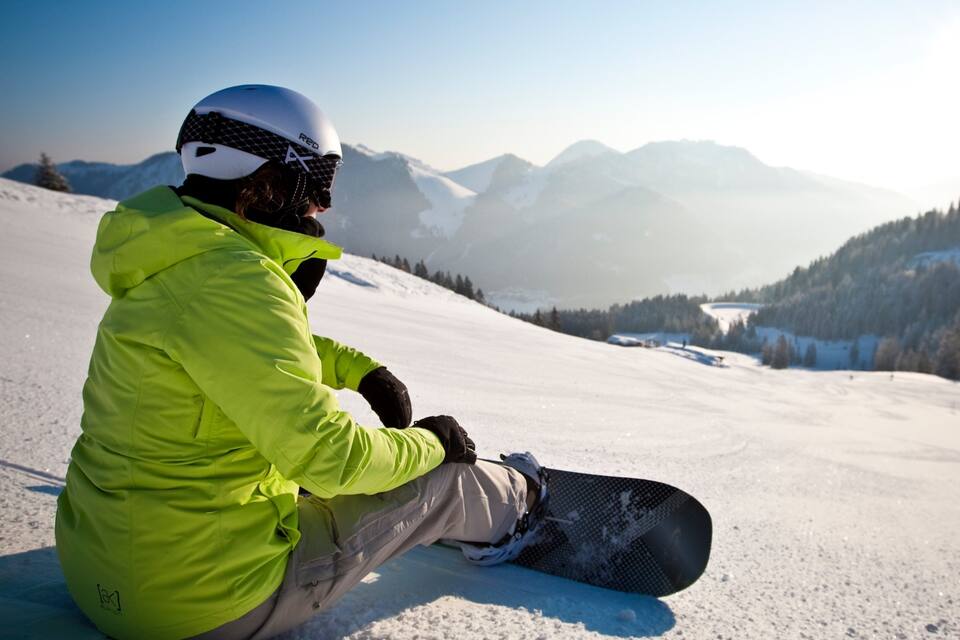 Een persoon zit op een met sneeuw bedekte helling met een snowboard, gekleed in een felgroene jas en een helm, met een bergachtig landschap op de achtergrond.