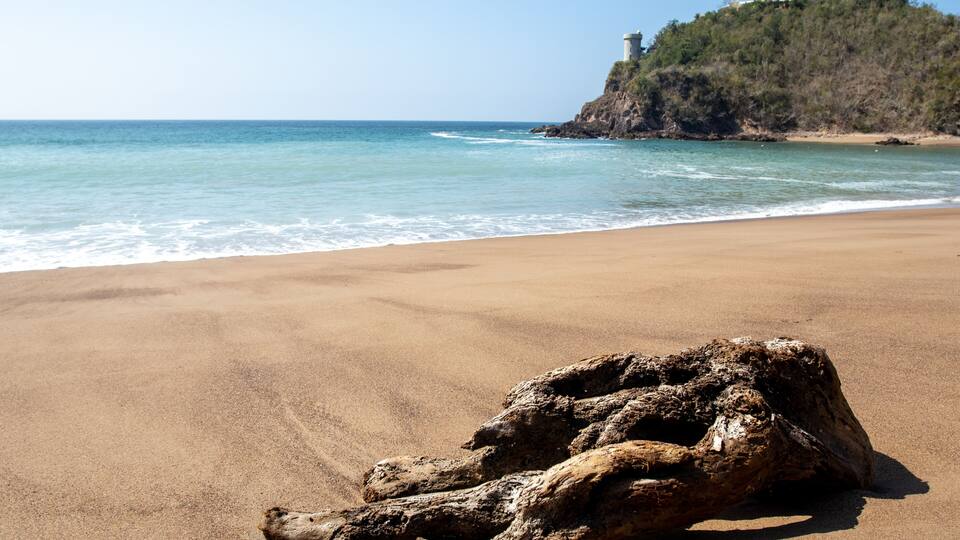 A sandy beach with a large rock formation and a lighthouse on a hill in the background.