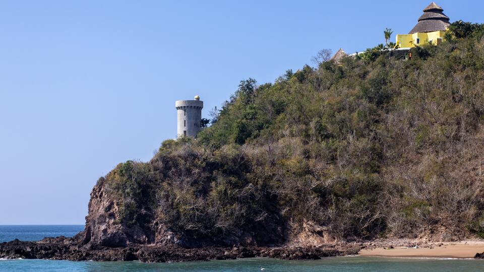 A coastal landscape with a lighthouse, a hilltop building, and a beach.