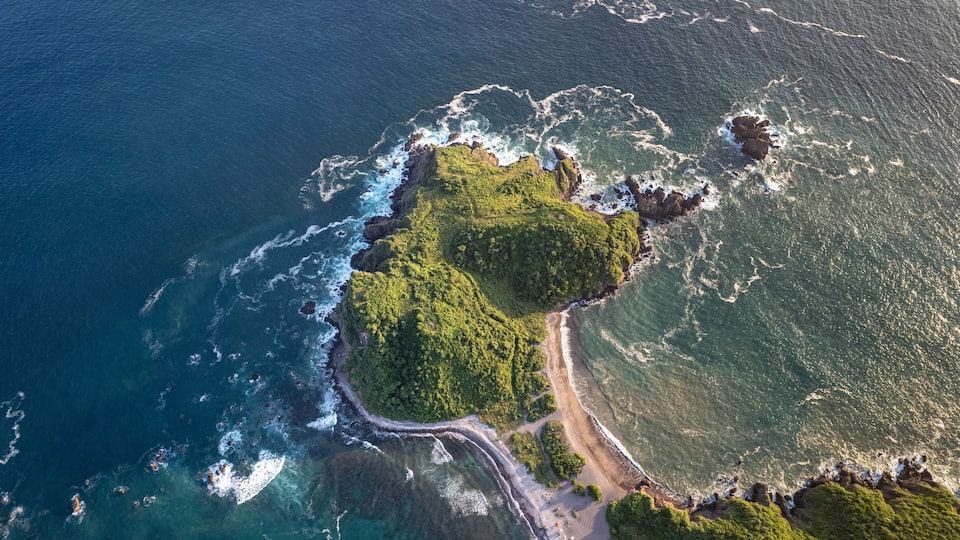 An aerial view of a coastal area with a narrow road winding along the shore, surrounded by lush green vegetation and rocky outcrops.