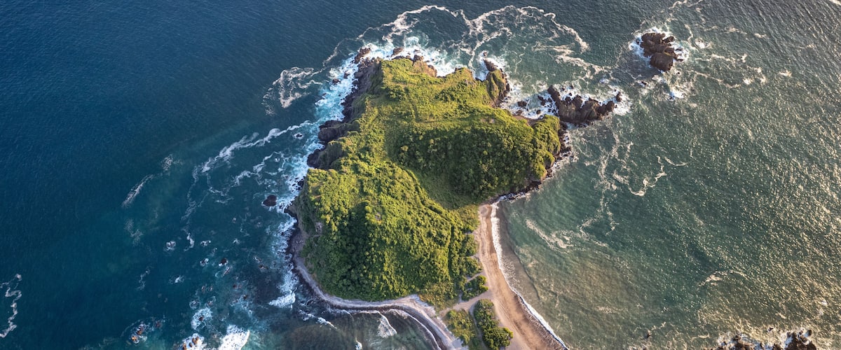An aerial view of a coastal area with a narrow road winding along the shore, surrounded by lush green vegetation and rocky outcrops.