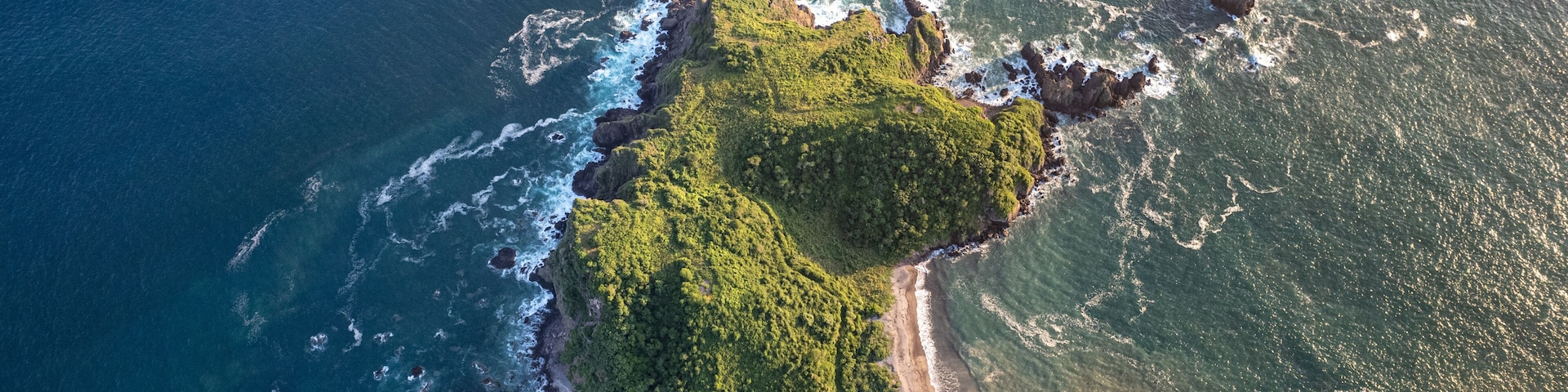 An aerial view of a coastal area with a narrow road winding along the shore, surrounded by lush green vegetation and rocky outcrops.