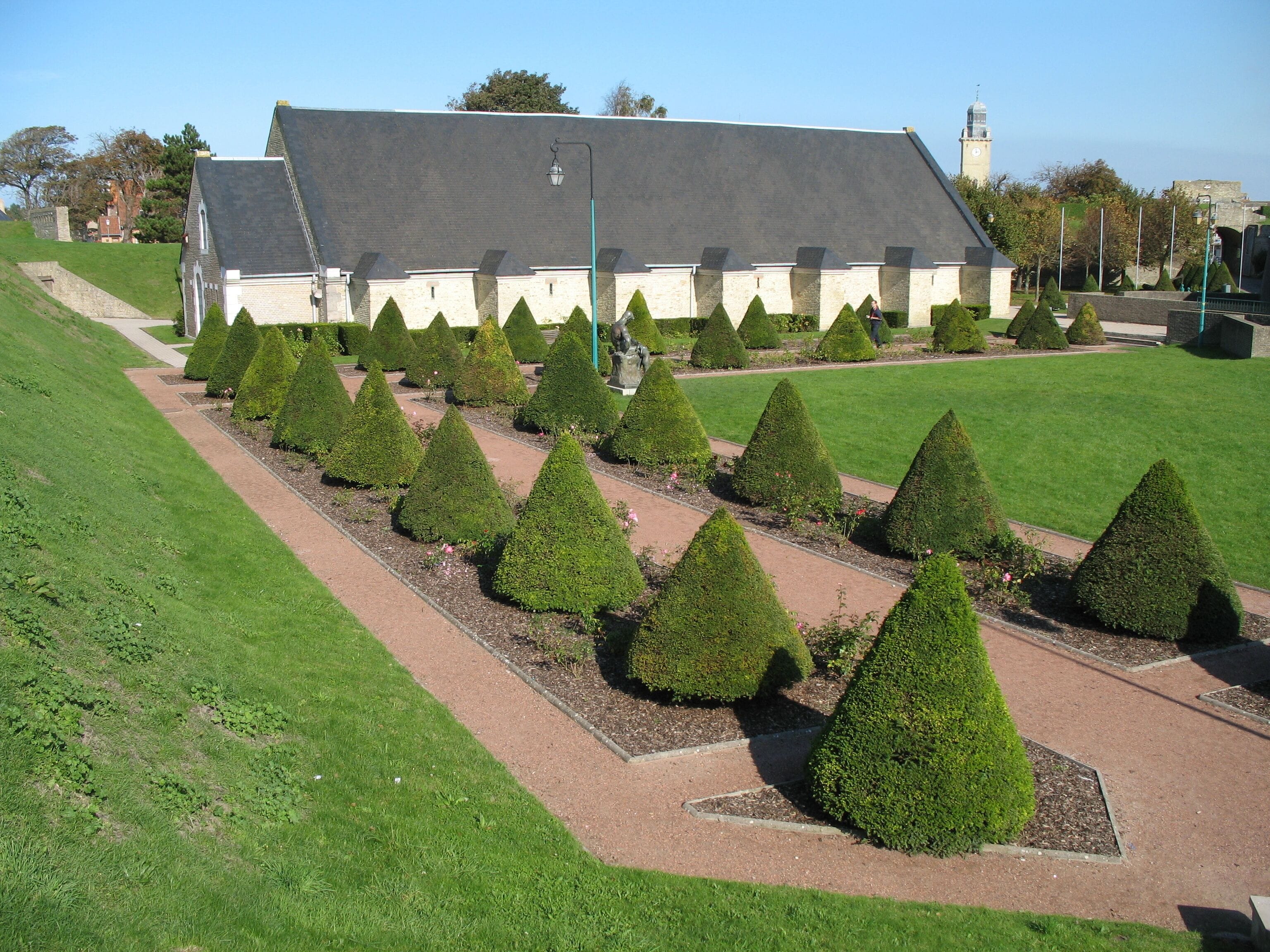 A building with a sloped roof, surrounded by a neatly trimmed garden with triangular hedges.