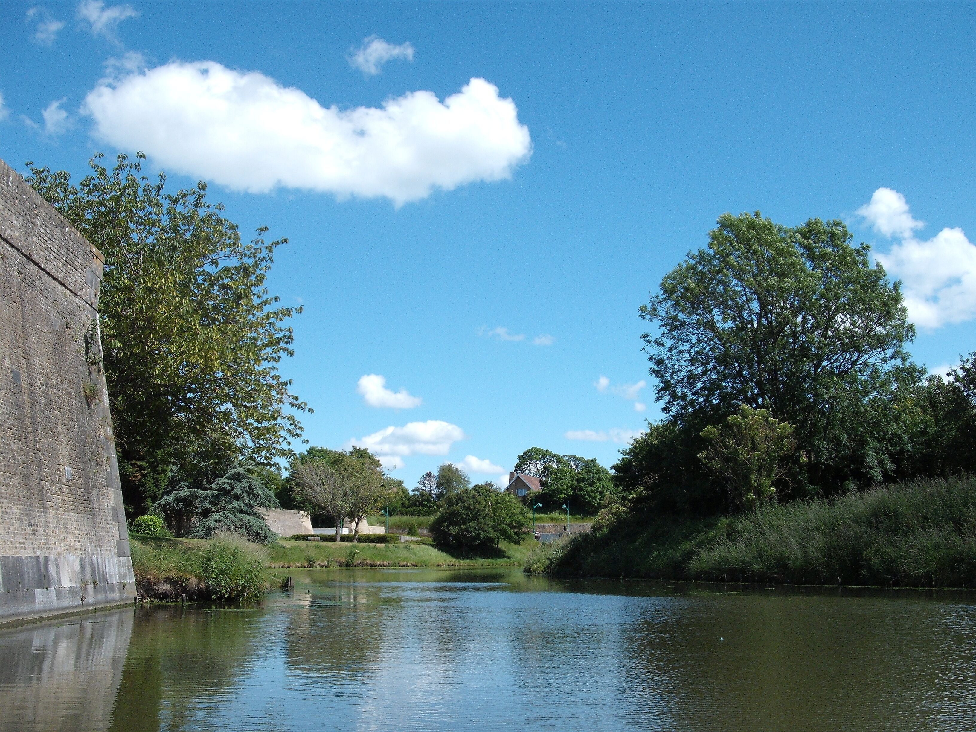 A river with a brick wall on the left, surrounded by trees and greenery.