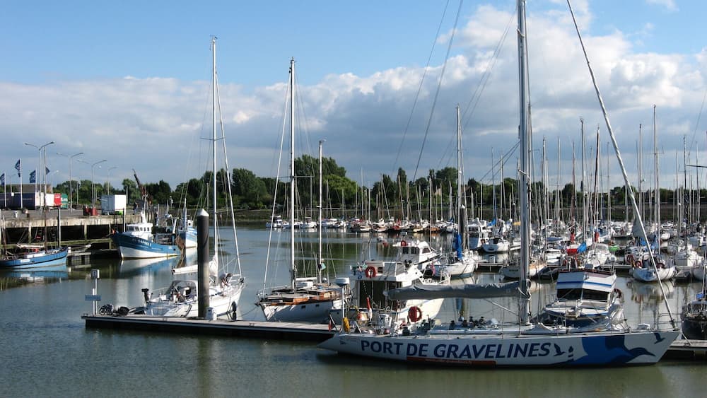 Un port de plaisance où sont amarrés de nombreux voiliers, un panneau bien visible indiquant « Port de Gravelines », et une étendue d’eau calme.