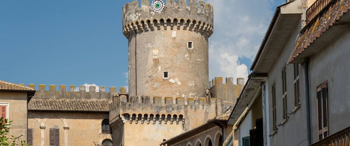 A historic tower with a crenellated top, a balcony with a railing, and a building with a tiled roof in the foreground.