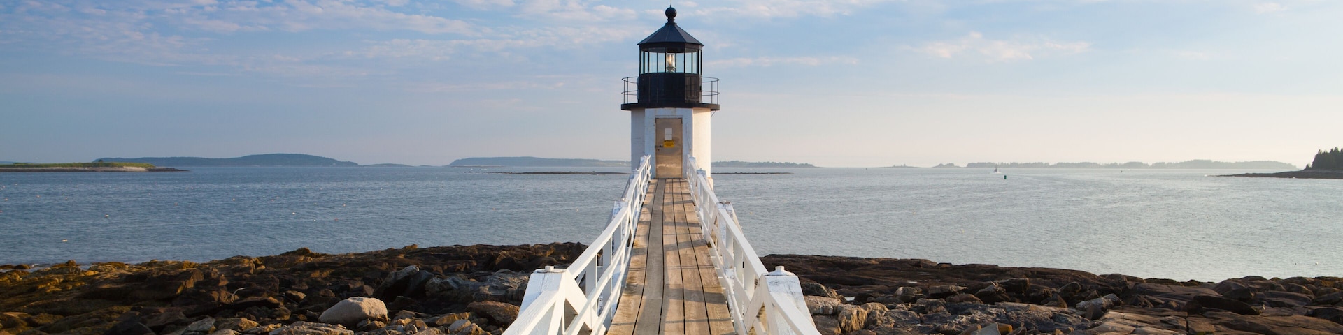 A wooden pier leading to a lighthouse by a rocky shoreline with calm water and distant land.