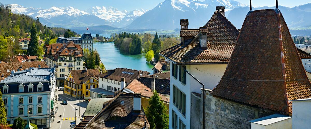 A town with historic buildings, a river, and snow-capped mountains in the background.