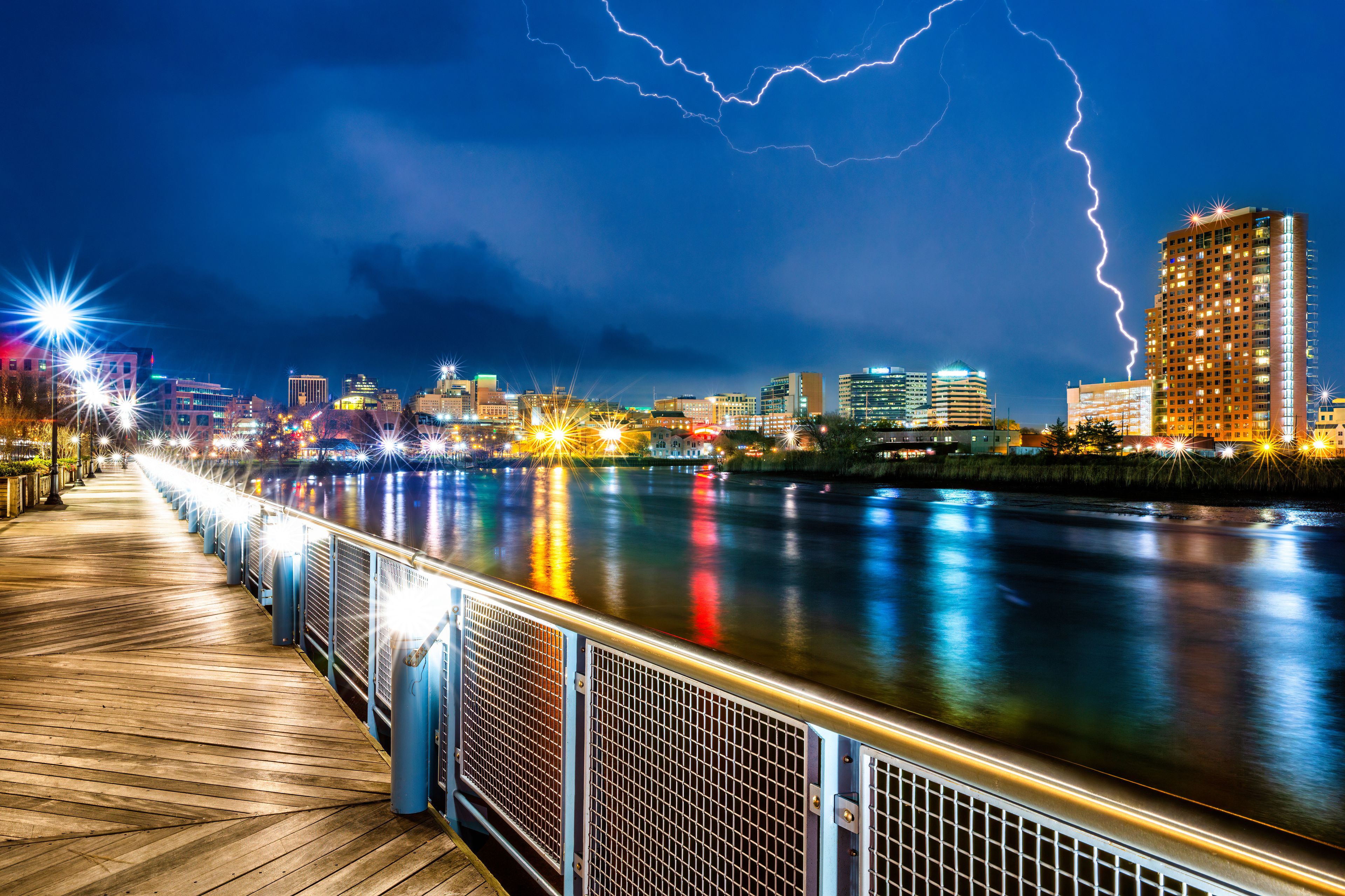 A waterfront cityscape at night with a wooden boardwalk, illuminated buildings, and lightning in the sky.