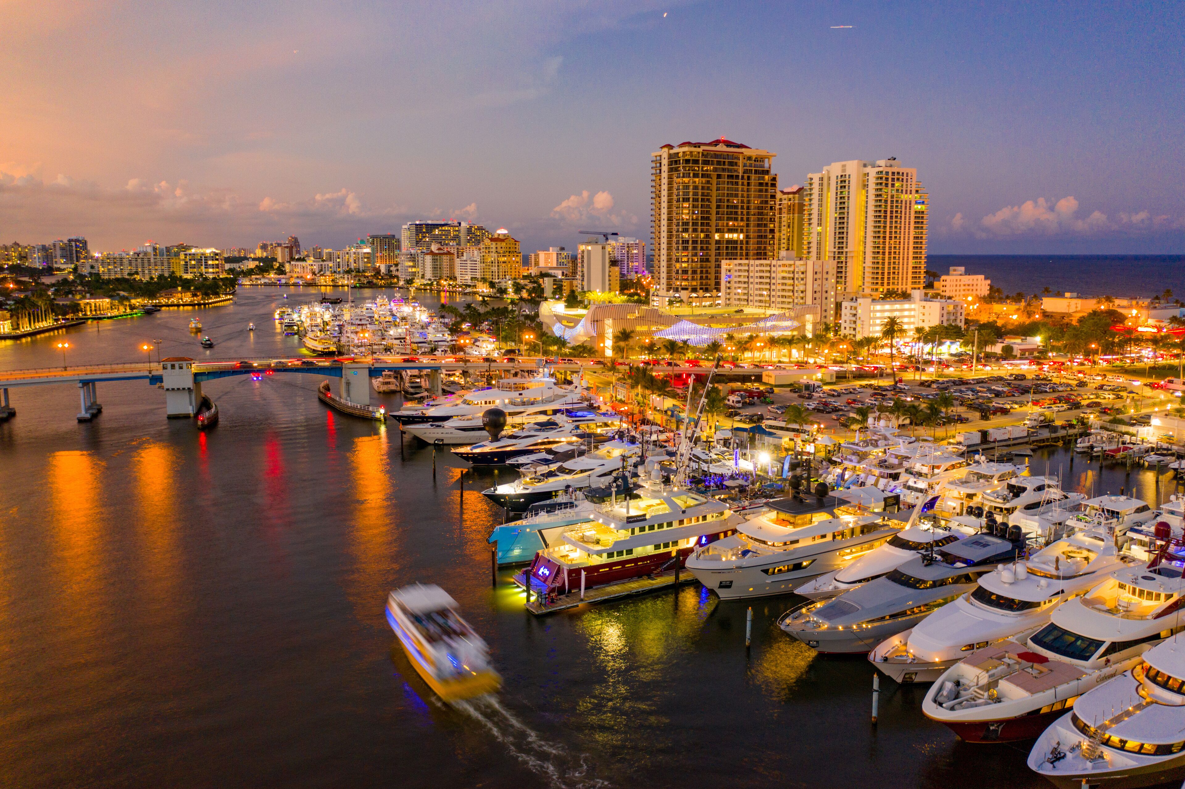 A marina with numerous boats docked, a bridge, and a city skyline with high-rise buildings.