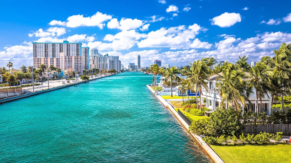 A waterfront cityscape with a turquoise river, tall buildings, and palm trees.