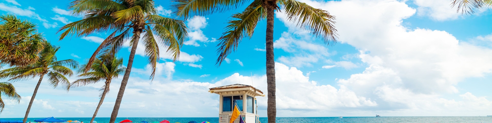 A beach with palm trees, a lifeguard tower, and people relaxing under umbrellas.