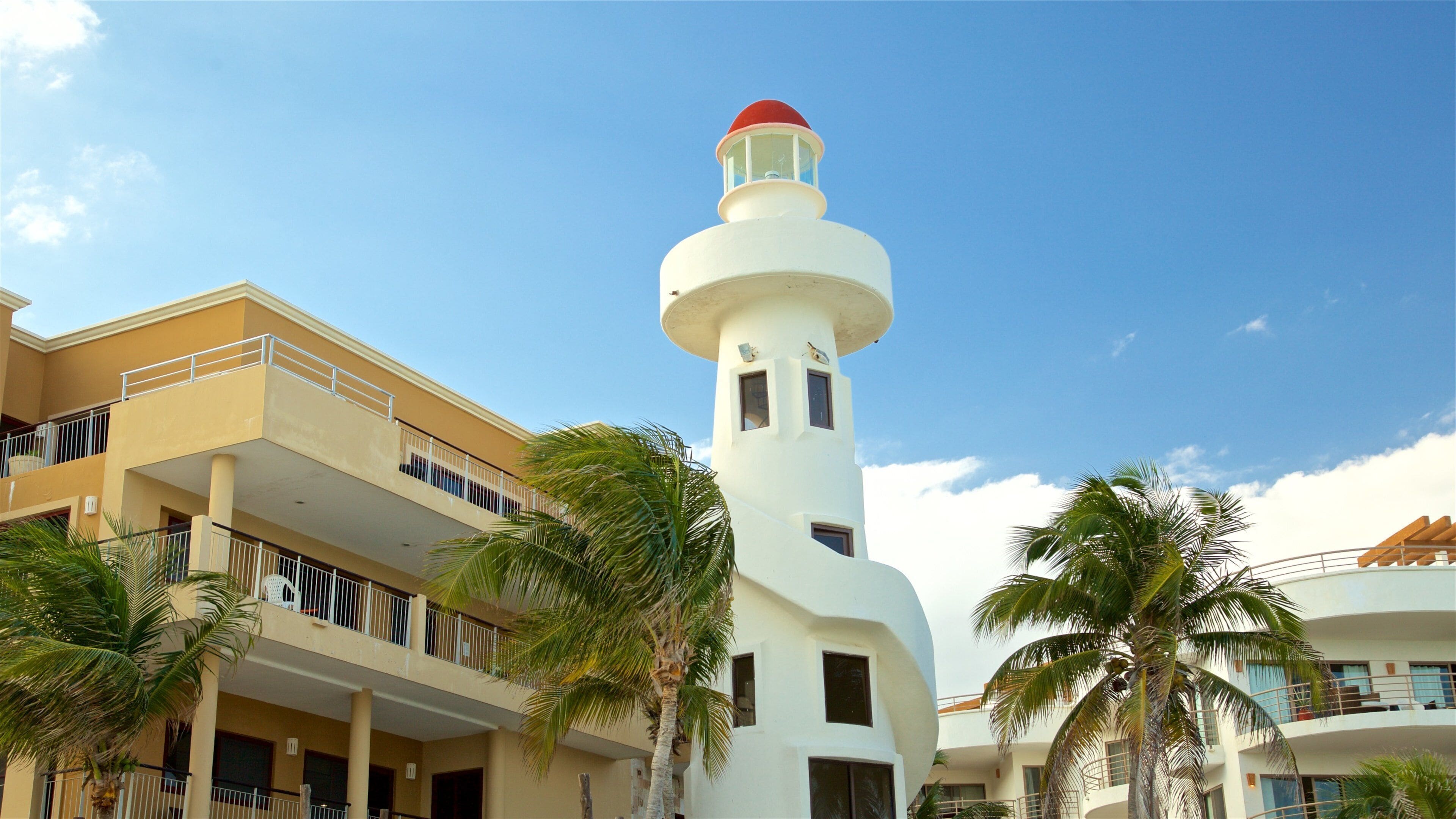 Un phare blanc coiffé d’un dôme rouge, entouré de palmiers et de bâtiments modernes.