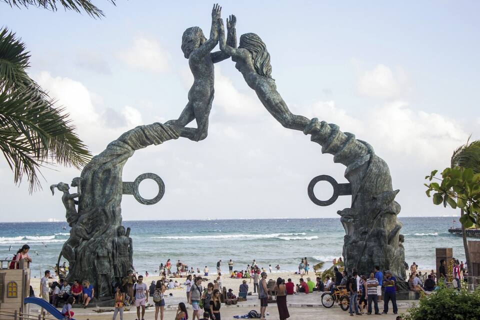 A beach with a large sculpture of two figures reaching out to each other, surrounded by palm trees and people.