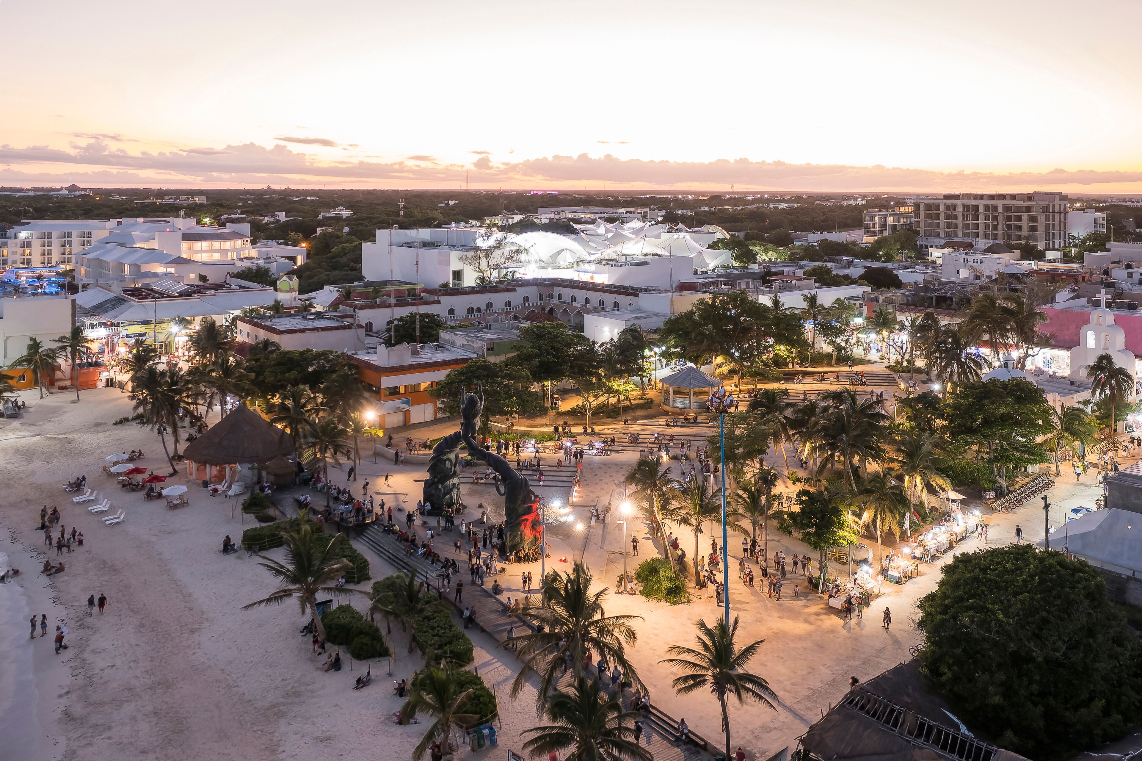 A coastal cityscape at sunset with a prominent beach area, palm trees, and a large white building with a dome.