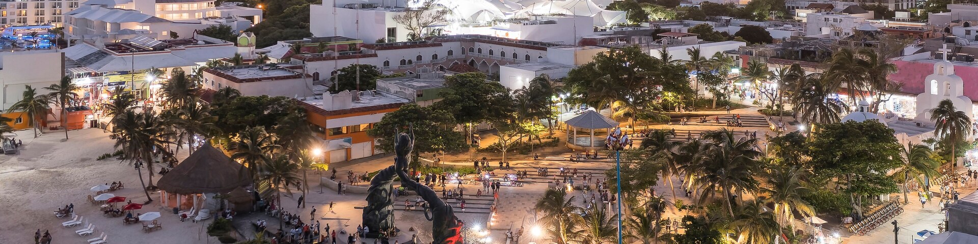Un paisaje urbano costero al atardecer, con una playa destacada, palmeras y un gran edificio blanco con una cúpula.