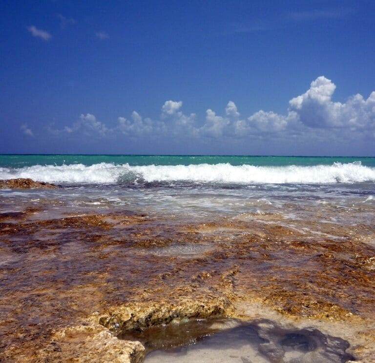 Uma praia com águas turquesa cristalinas, margem rochosa e céu claro com nuvens.