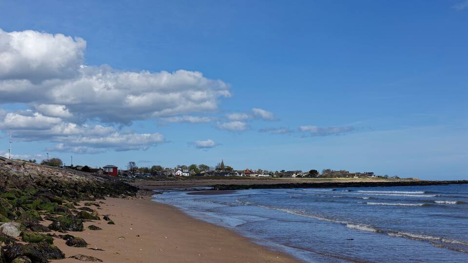 A sandy beach with waves, a rocky shoreline, and houses in the background.