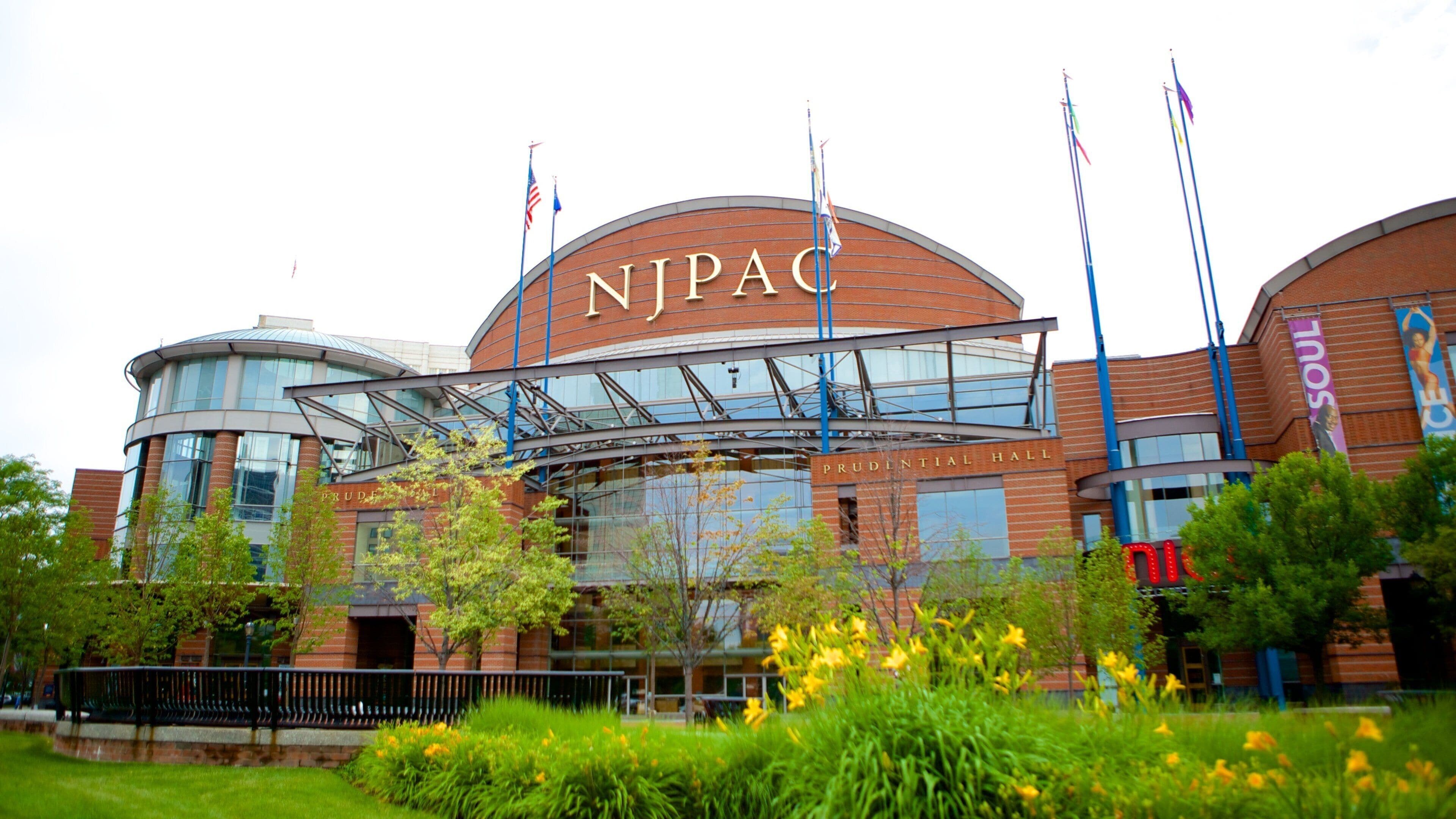 Een modern gebouw met een gebogen gevel, waarop het bord “NJPAC” duidelijk zichtbaar is, en meerdere vlaggen op vlaggenmasten.