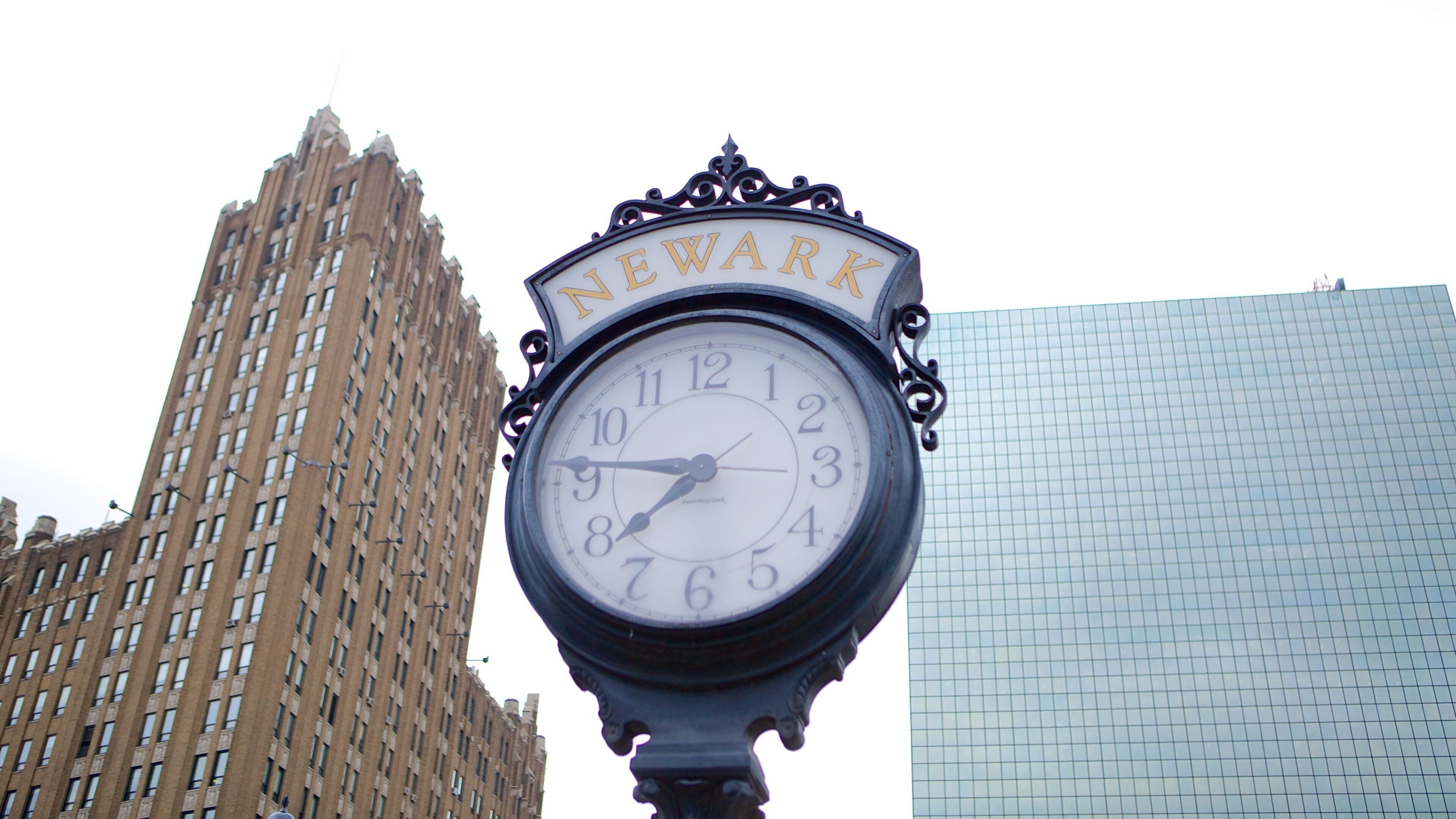 A clock with 'NEWARK' written on it, set against a tall building and a modern glass structure.