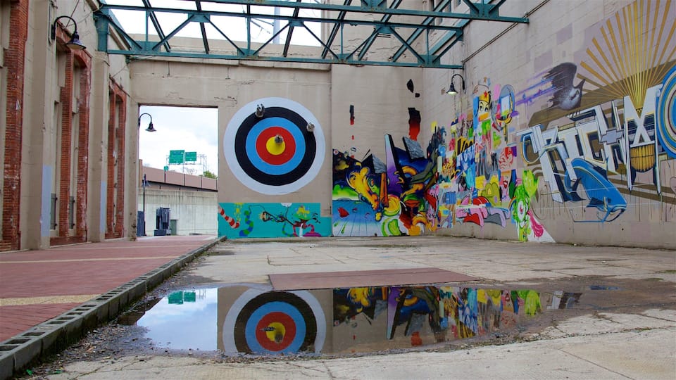 Uma passagem urbana com arte de graffiti, um grande mural em forma de alvo e uma poça refletindo a cena.