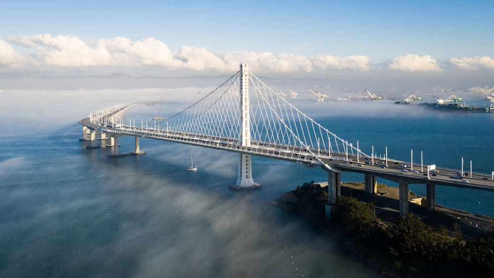 A cable-stayed bridge over a body of water with a lighthouse and ships in the distance.