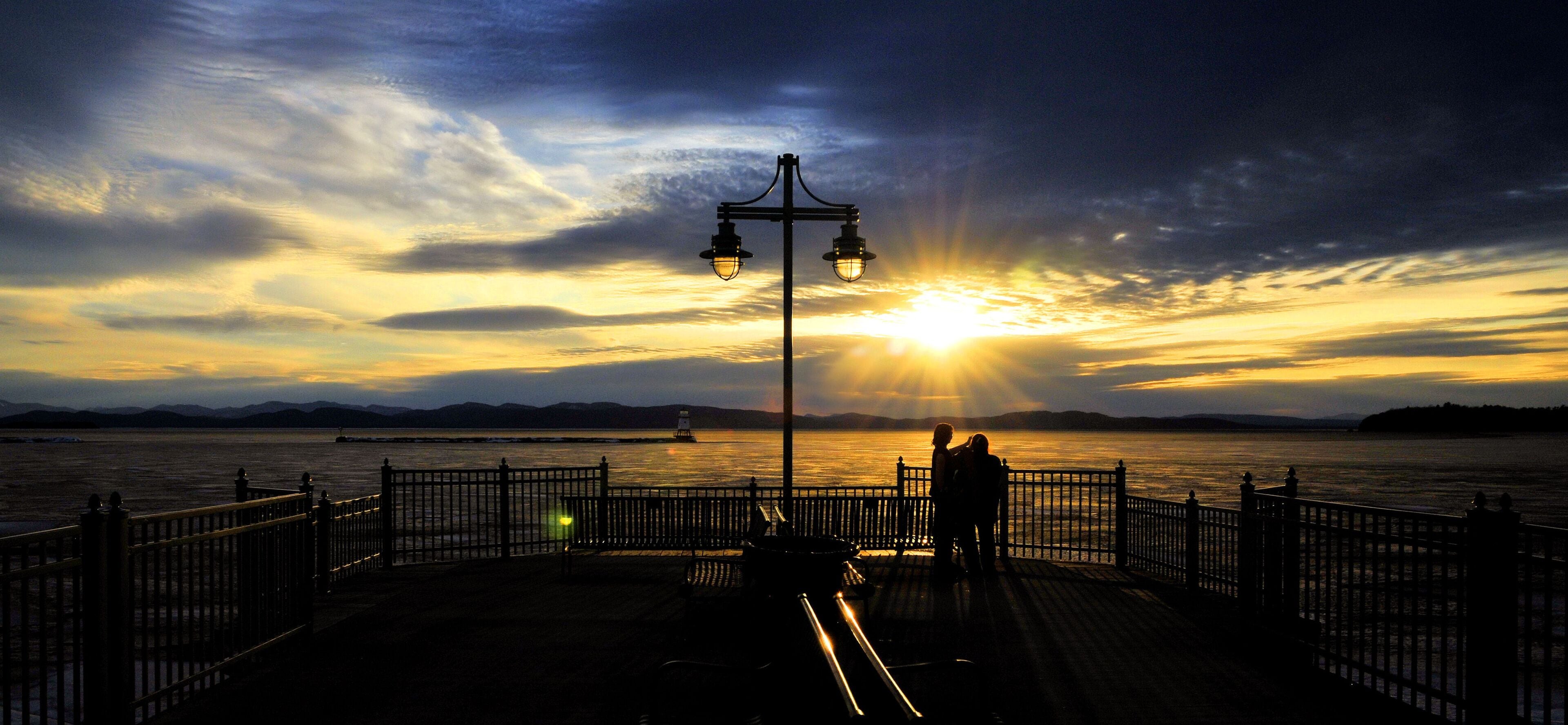 A sunset over a body of water with a pier, a lamp post, and distant mountains.