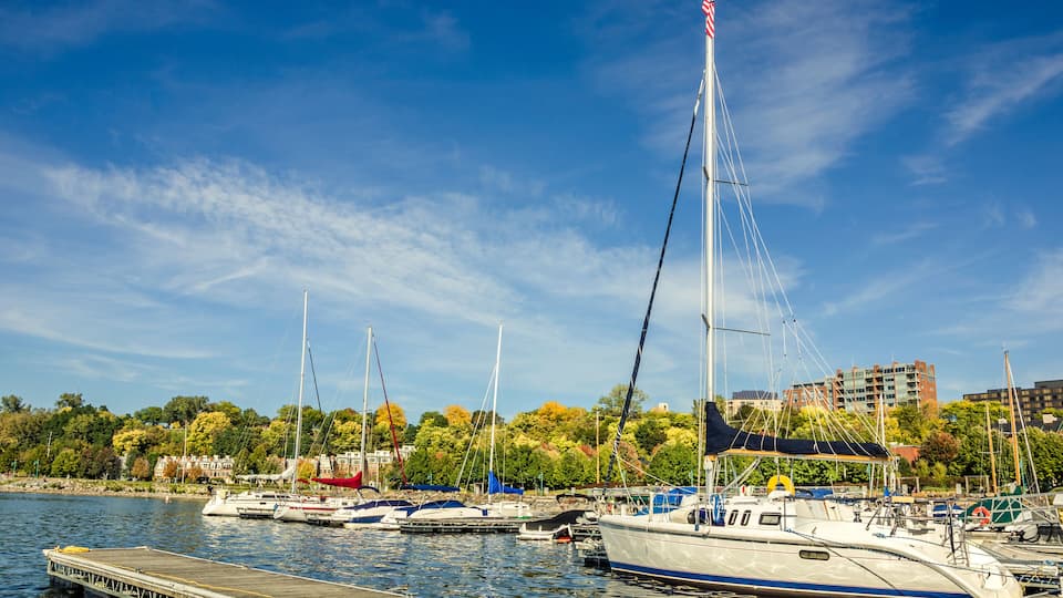 A marina with several sailboats docked at a wooden pier, surrounded by autumn-colored trees and buildings in the background.
