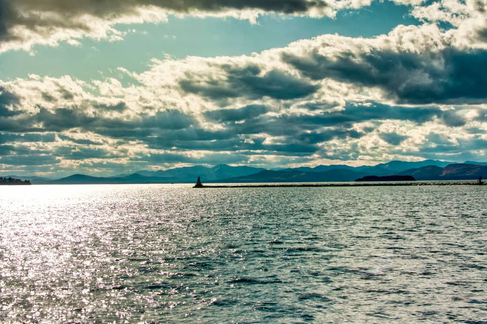 Massa de água com um barco, céu claro com nuvens dispersas e montanhas ao longe.