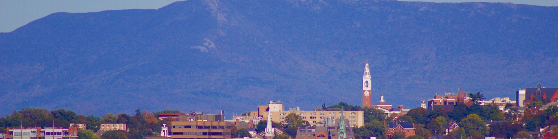 Un paysage urbain côtier avec un clocher d’église imposant, une montagne en arrière-plan et un mélange de bâtiments modernes et historiques.