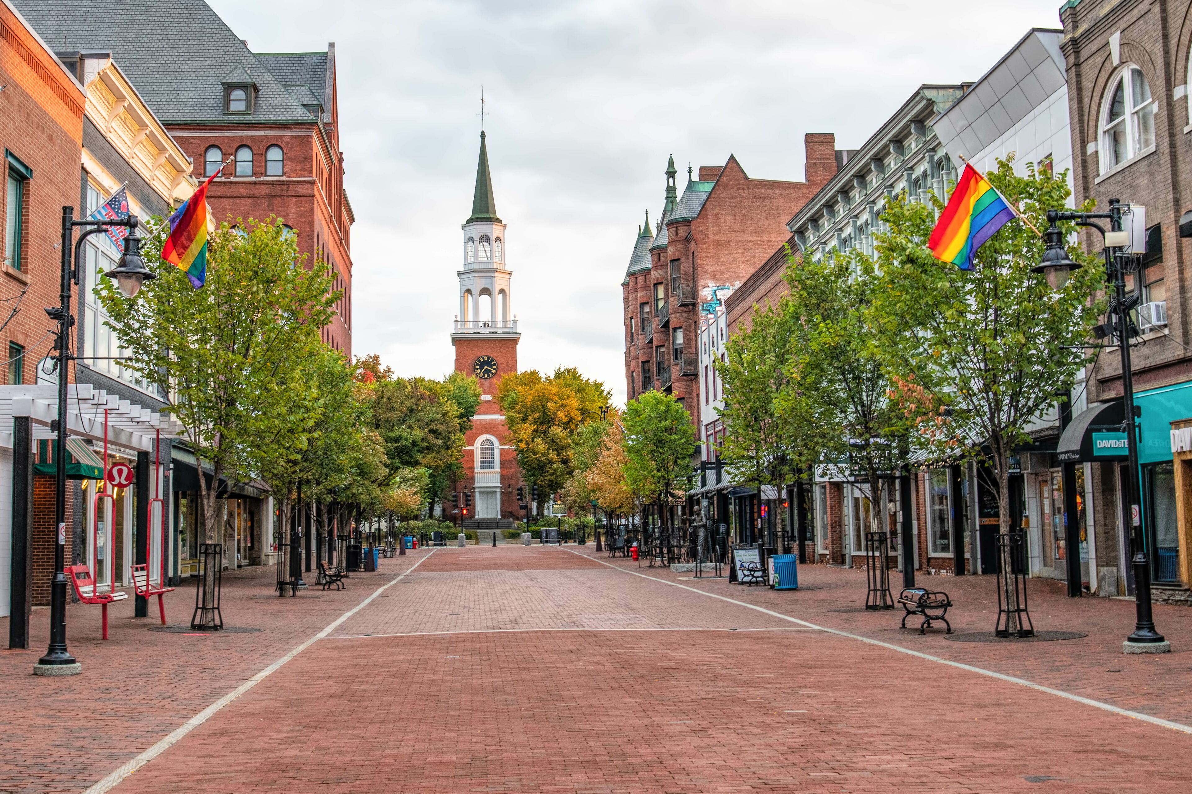 A brick-paved street lined with trees and buildings, a church steeple, and rainbow flags.
